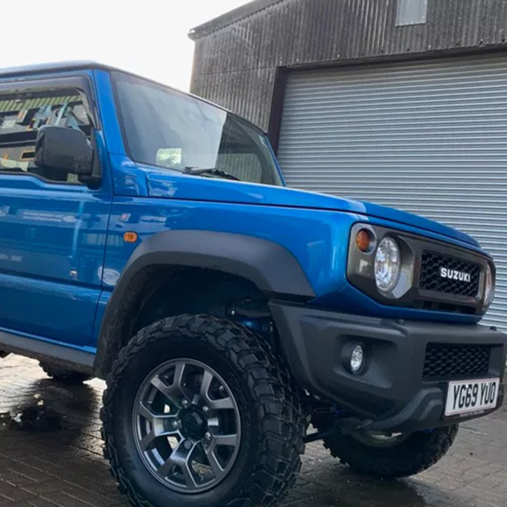 A blue Suzuki Jimny parked in front of a garage, equipped with the ARB Old Man Emu Front Nitrocharger Sport 60154 for Suzuki Jimny (4th Gen) 4 Cylinder 1.5L Petrol Engine All Models by Old Man Emu for optimal performance.