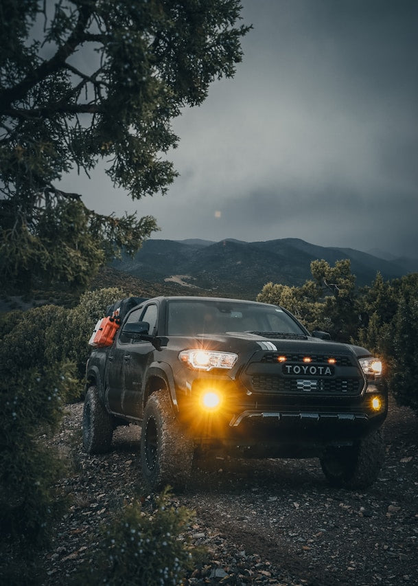 Toyota Tacoma with off-road tires, lifted suspension, front LED lights, and fuel cans mounted on the bed off-roading at dusk