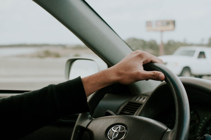 Driver's hand gripping Toyota steering wheel with dashboard and side mirror visible on a clear day