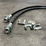 A pair of Mudify Extended Front Brake Lines for Toyota 4Runner 2003-ON, FJ Cruiser 2007-ON, Lexus GX460 & GX470 2003-ON stainless steel brake hoses on a concrete surface.