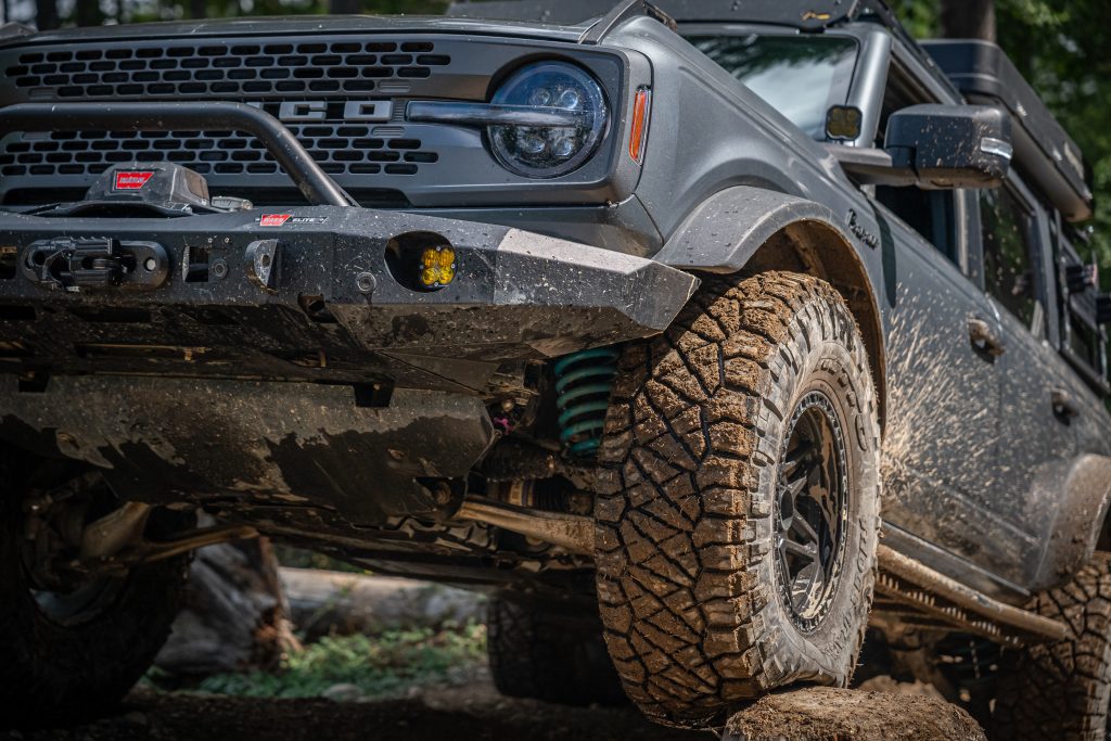 Gray Ford Bronco off road SUV with mud covered all terrain tire, front bumper, and visible suspension on rocky terrain
