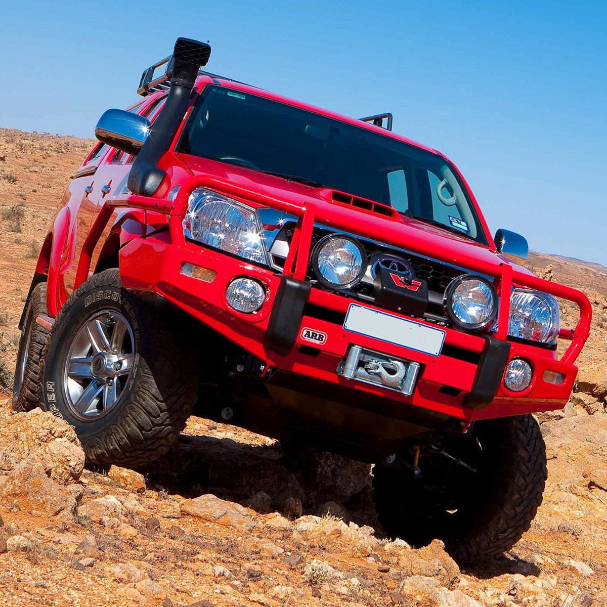 An OME 2.5 - 3 inch Essentials Lift Kit Hilux Vigo (05-15)-equipped red Toyota Tacoma is driving through a desert, showcasing its exceptional ground clearance and advanced suspension system.