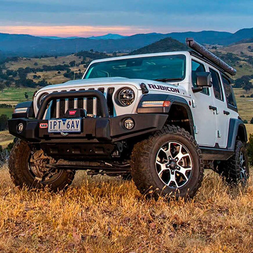 A white Old Man Emu Jeep Wrangler JL 4 Door (18-23) with impressive ground clearance parked in a field with mountains in the background.