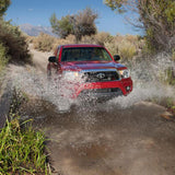 A red Toyota Tacoma with an Old Man Emu OME 2.5 inch Lift Kit for Tacoma (05-15) - Front Shocks Assembly is driving down a dirt road, demonstrating impressive ground clearance and suspension articulation.