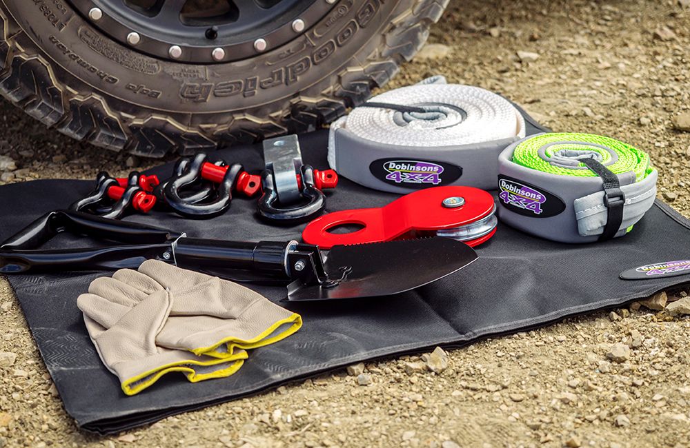 Off road recovery gear with shackles straps shovel gloves and an all terrain tire on a ground mat