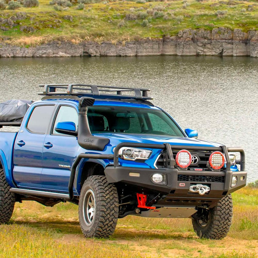 A blue Old Man Emu Toyota Tacoma with ARB Old Man Emu Upper Control Arms UCA0005 is parked near a lake.