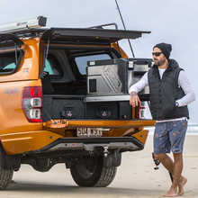 Load image into Gallery viewer, A man standing next to an ARB Elements 63Quart Portable Fridge Freezer Weatherproof Stainless Steel 10810602 SUV on the beach.