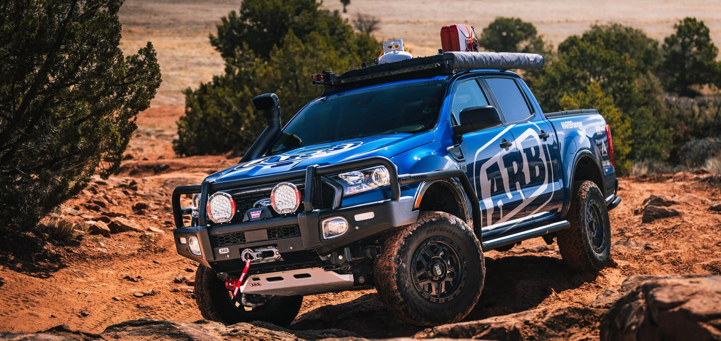 An ARB blue pickup truck is driving through a rocky area, overcoming the challenging terrain with its steel frame and ARB Rated LHS Recovery Point for Toyota Hilux 2814020.