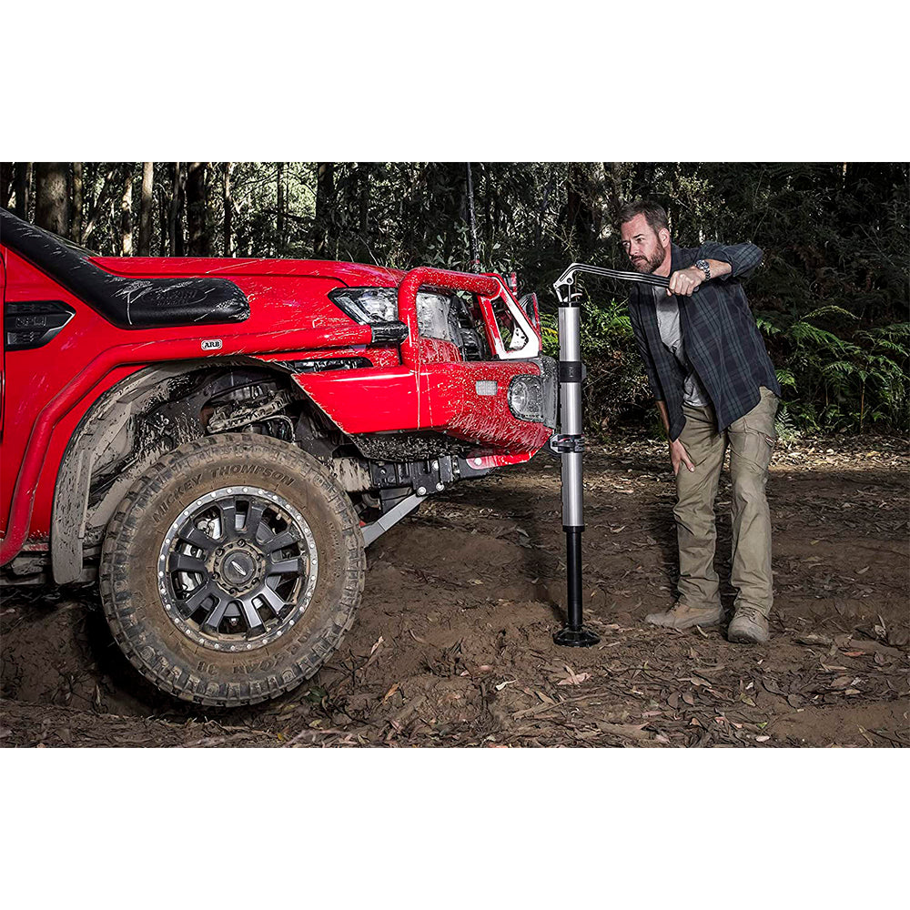 A man operating an ARB Hydraulic Recovery Jack 1060001 lowering lever next to a red truck in the dirt, utilizing hydraulic technology for recovery missions.