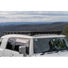 Load image into Gallery viewer, A vehicle-specific Toyota FJ Cruiser parked on a hill, equipped with an ARB Base Rack Deflector (17920040) for extra storage.
