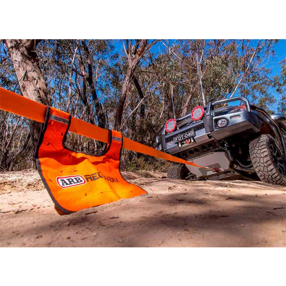 An orange strap is securely attached to a 4-wheel drive jeep on a dirt road, emphasizing safety precautions and the use of an ARB Recovery Winch Damper ARB220.