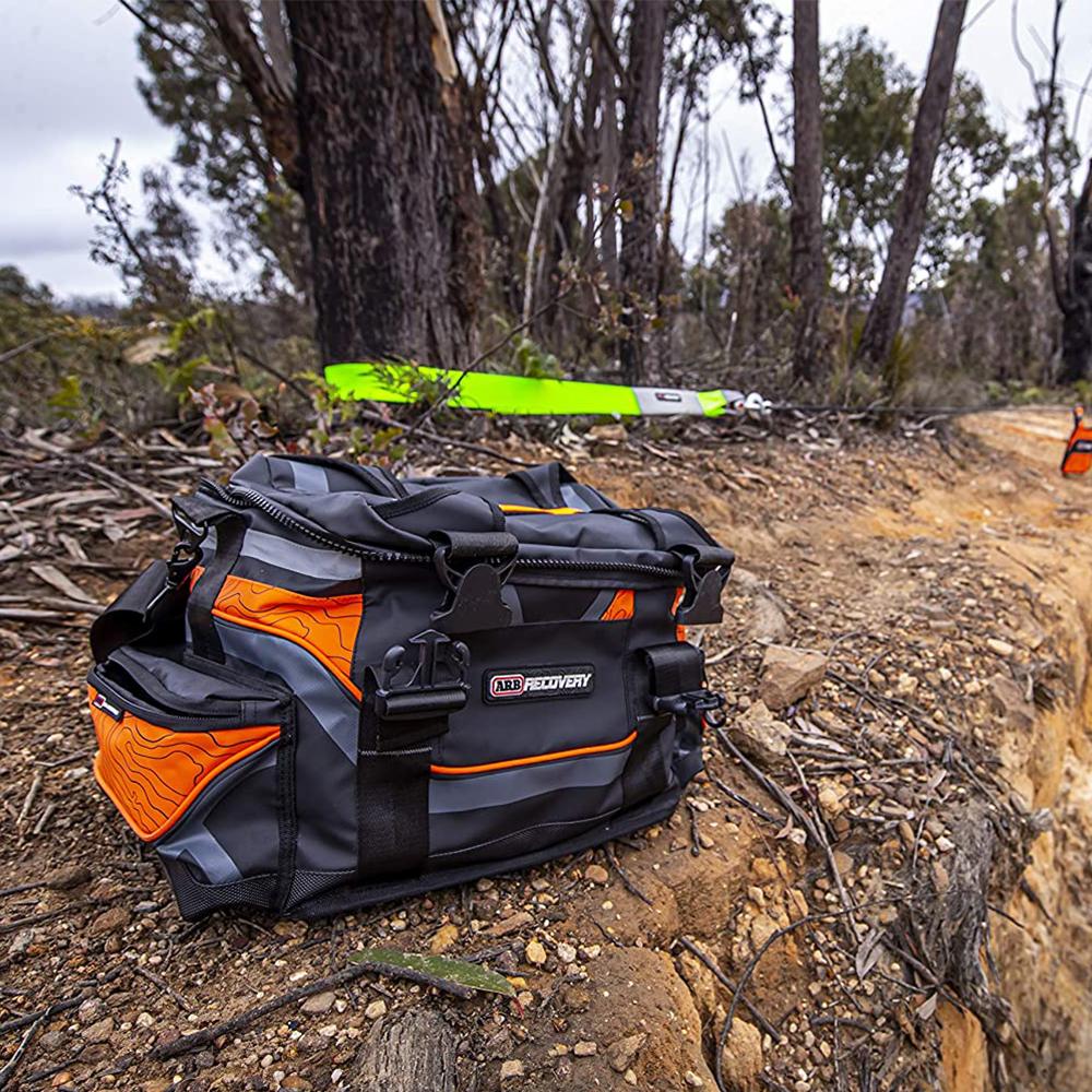 An ARB Orange Large Recovery Bag ARB501A sitting on a dirt trail, filled with vehicle recovery gear such as a vehicle snatch strap from the ARB Premium Recovery Kit.