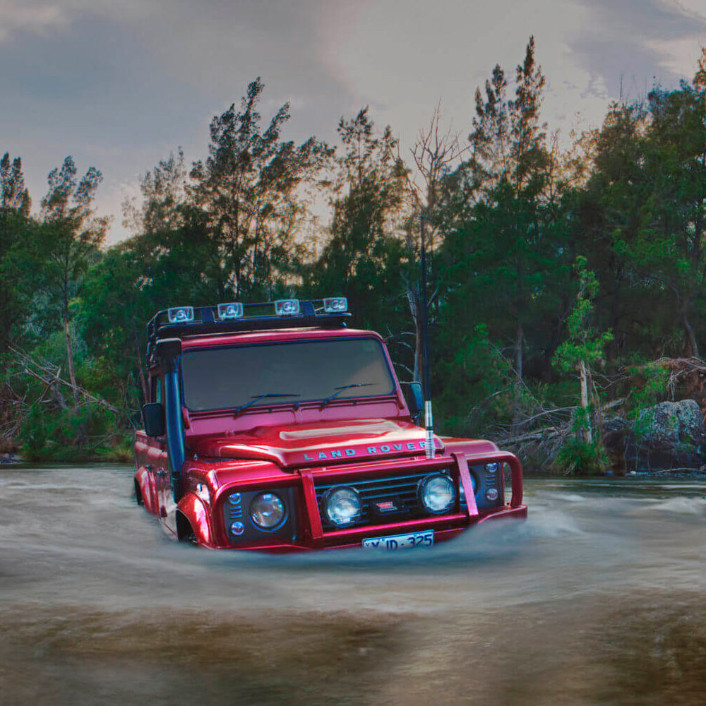 A robust red Old Man Emu Land Rover effortlessly maneuvers through a watery terrain, showcasing its exceptional ground clearance with the OME 2 inch Lift Kit for Land Rover Defender 110 (85-17).