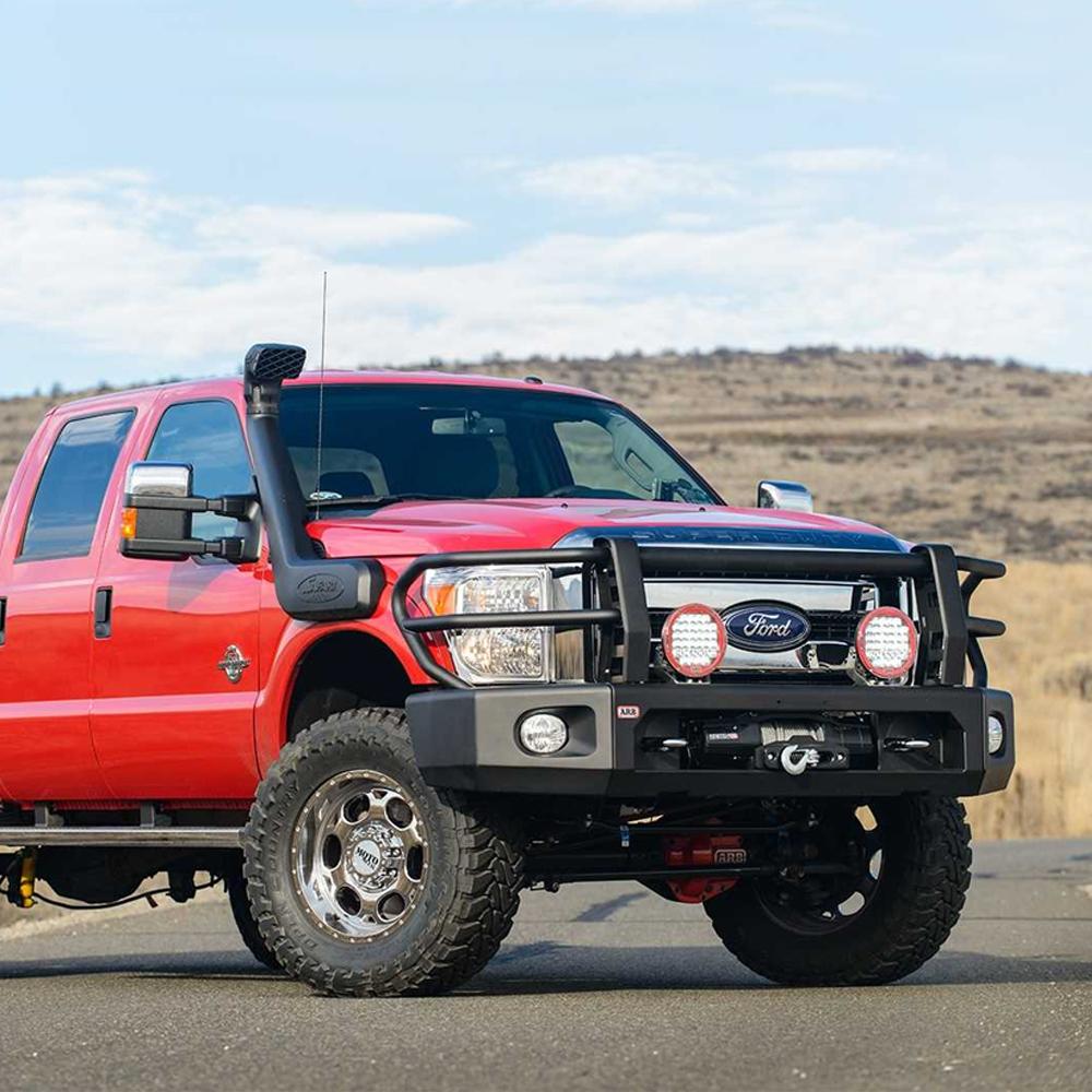 A red ARB Kit Textured Modularbar Type A for FORD F-250 SUPER DUTY (2011 - 2016) truck is parked on the side of a road, boasting LED indicators.