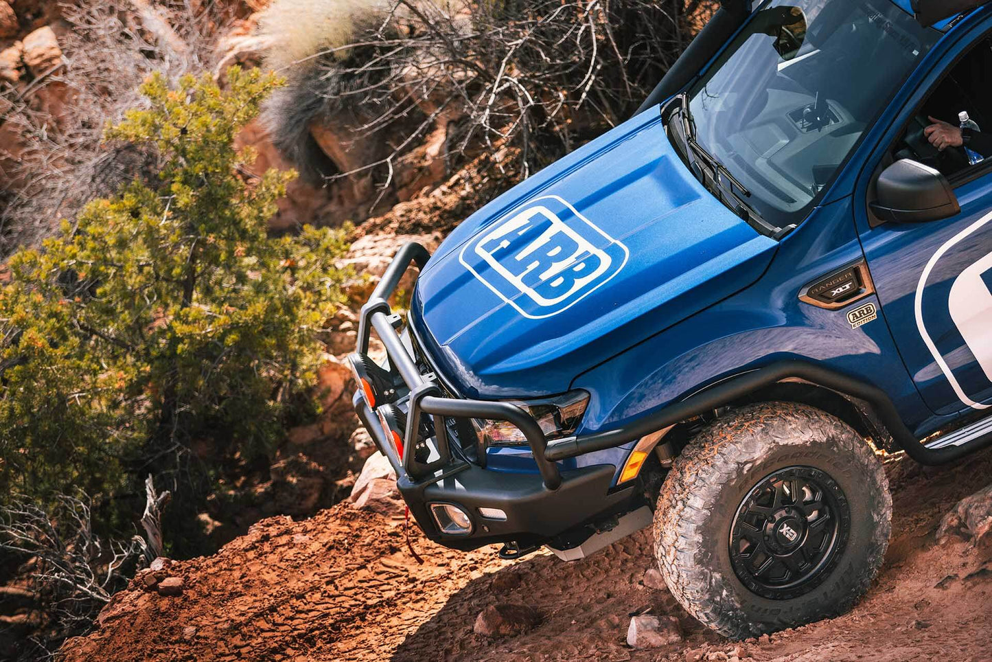 A blue Toyota Hilux Revo/Rocco is driving down a rocky trail, equipped with the ARB Sahara Front Top Tube For Toyota Hilux Revo/Rocco (2018-2022) 5100350 bumper kit from ARB.