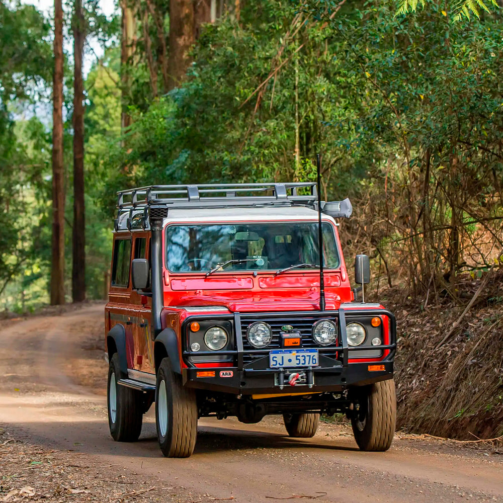 An OME 2 inch Lift Kit for Land Rover Defender 110 (85-17) equipped red Land Rover is confidently driving down a dirt road, gliding effortlessly over the uneven terrain thanks to its exceptional ground clearance and advanced suspension system.