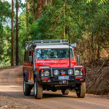 Load image into Gallery viewer, An OME 2 inch Lift Kit for Land Rover Defender 110 (85-17) equipped red Land Rover is confidently driving down a dirt road, gliding effortlessly over the uneven terrain thanks to its exceptional ground clearance and advanced suspension system.