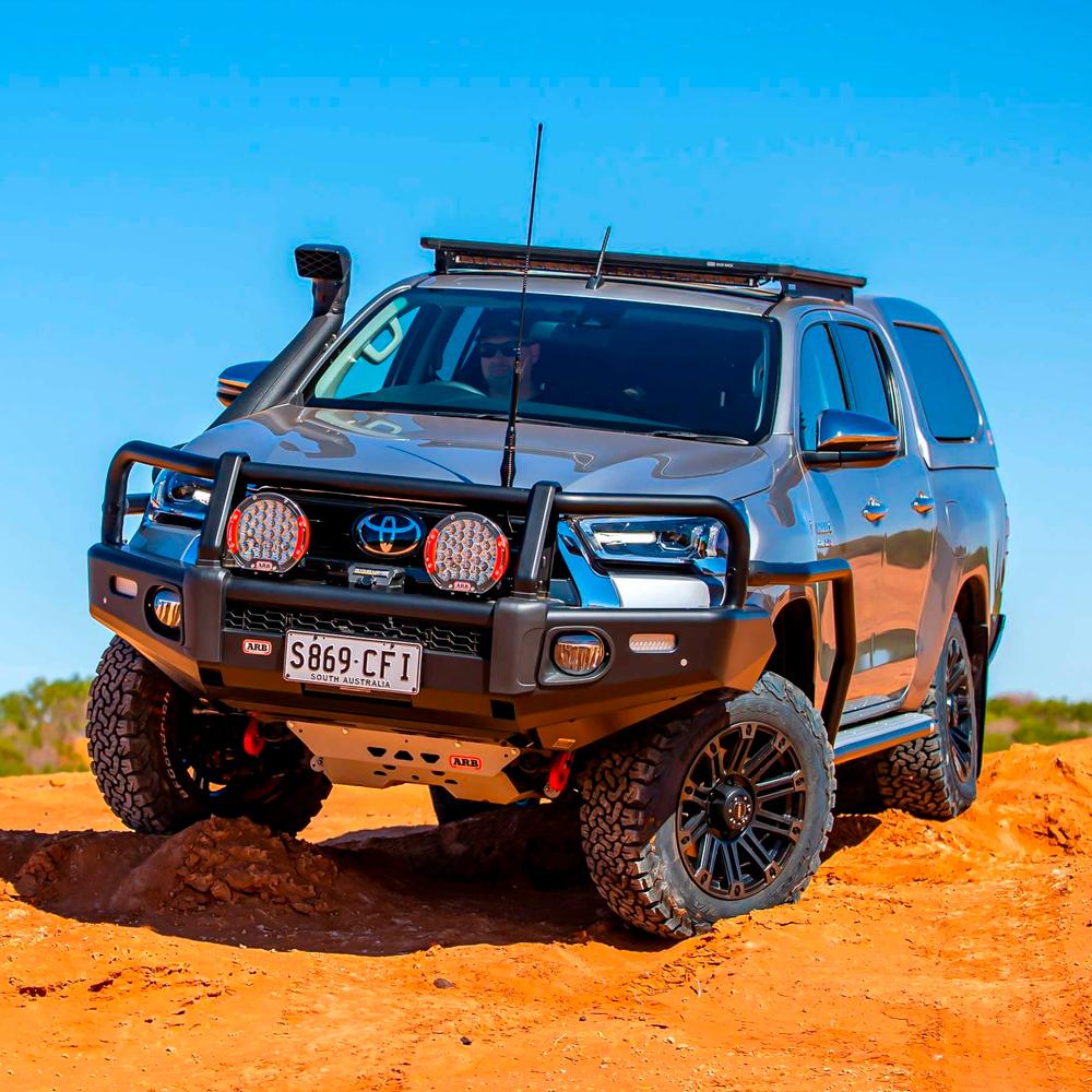 The ARB Alloy Flat Rack 70” X 44” with Mesh Floor for Toyota Land Cruiser 100 Series 1997-2007, equipped with a fitting kit, is driving on a dirt road.
