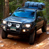 A Toyota Tacoma with an ARB Summit Bumper is driving down a trail in the woods, protected by Zinc Rich Primer and equipped with an ARB Roller Fairlead Cover.