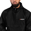 A man with a beard is wearing the Mudify Embroidered Mudify Packable Jacket, a wind and rain resistant black jacket with the word muddy on it.