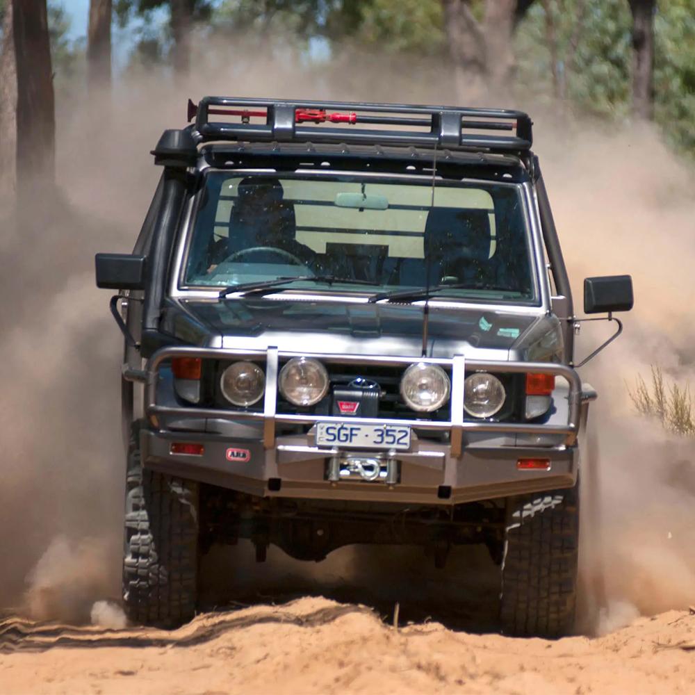 An ARB off-road vehicle equipped with the Steel Roof Rack Basket with Mesh Floor for Toyota ARB 3813020M drives through a dense forest, navigating rugged terrain with the help of a fitting kit for added stability. The vehicle effortlessly crosses over a natural drainage channel without.