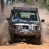 An ARB off-road vehicle equipped with the Steel Roof Rack Basket with Mesh Floor for Toyota ARB 3813020M drives through a dense forest, navigating rugged terrain with the help of a fitting kit for added stability. The vehicle effortlessly crosses over a natural drainage channel without.