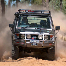 Load image into Gallery viewer, An ARB off-road vehicle equipped with the Steel Roof Rack Basket with Mesh Floor for Toyota ARB 3813020M drives through a dense forest, navigating rugged terrain with the help of a fitting kit for added stability. The vehicle effortlessly crosses over a natural drainage channel without.