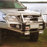 A silver Toyota Hilux with an ARB Deluxe Winch Front Bumper 3421600 for Toyota Fortuner 2006 - 2011 parked next to a body of water.
