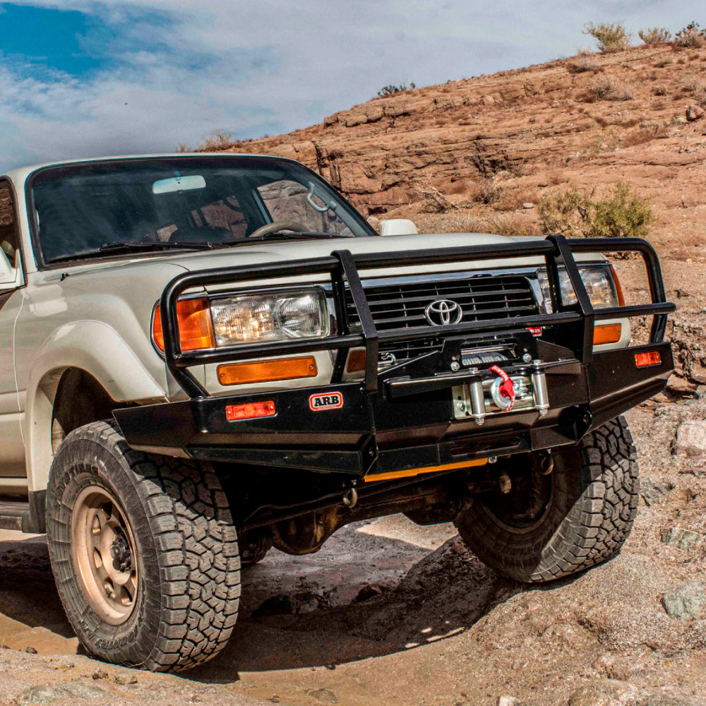 An ARB Deluxe Winch Front Bumper 3411050 for Land Cruiser 80 Series 1990-1997, known for its durable steel construction, is driving through a rocky area.