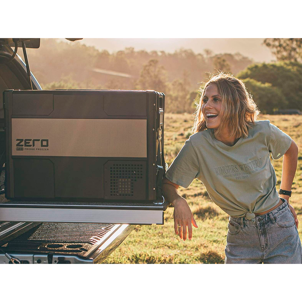 A woman standing next to an ARB Zero 73 Quart Dual Zone Portable Fridge Freezer 10802692 in the back of a truck.