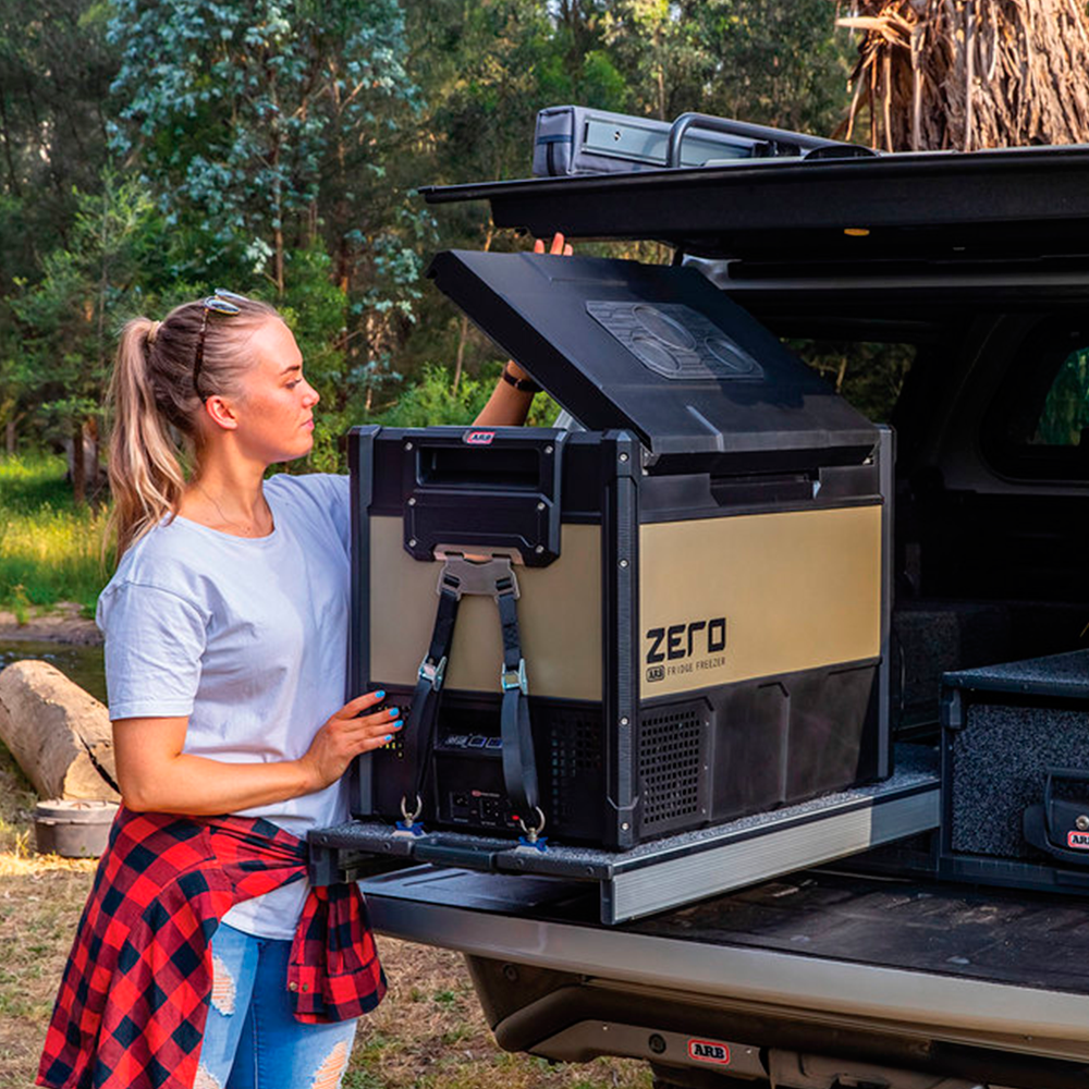 A woman securely placing an ARB Zero 63 Quart Single Zone Portable Fridge Freezer 10802602 in the back of a truck, ensuring maximum cooling capacity. She utilizes wireless monitoring to easily keep track of the temperature and maintain proper storage conditions during.