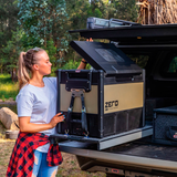 A woman securely placing an ARB Zero 63 Quart Single Zone Portable Fridge Freezer 10802602 in the back of a truck, ensuring maximum cooling capacity. She utilizes wireless monitoring to easily keep track of the temperature and maintain proper storage conditions during.