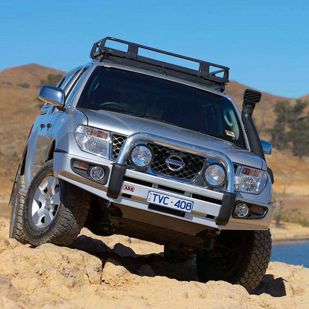 The ARB Roof Rack Fitting Kit for Nissan Pathfinder and NAVARA D40 3738010, equipped with secure storage and steel roof racks, is driving on sand near a body of water.