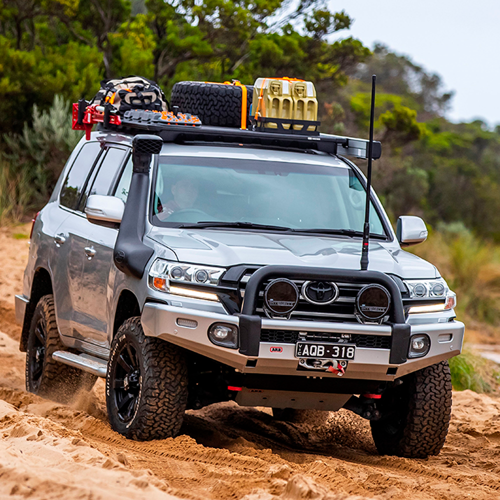 An ARB Alloy Flat Rack With Mesh Floor for Toyota Land Cruiser 200 Series ARB 4900040MKLC2 is driving down a dirt road, ensuring maximum protection and airbag compatibility while maintaining a suitable approach angle.