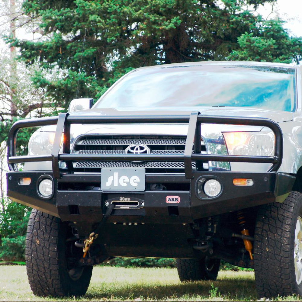 A Toyota Tacoma with durable steel construction parked in a field, featuring an "Deluxe Bull Bar Winch Bumper For Toyota Tundra 2007-2013 ARB 3415010" by ARB.