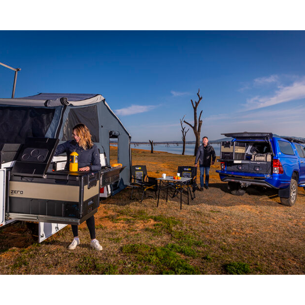 A woman is standing next to a durable and versatile ARB Zero 101 Quart Dual Zone Portable Fridge Freezer 10802962 blue camper trailer.