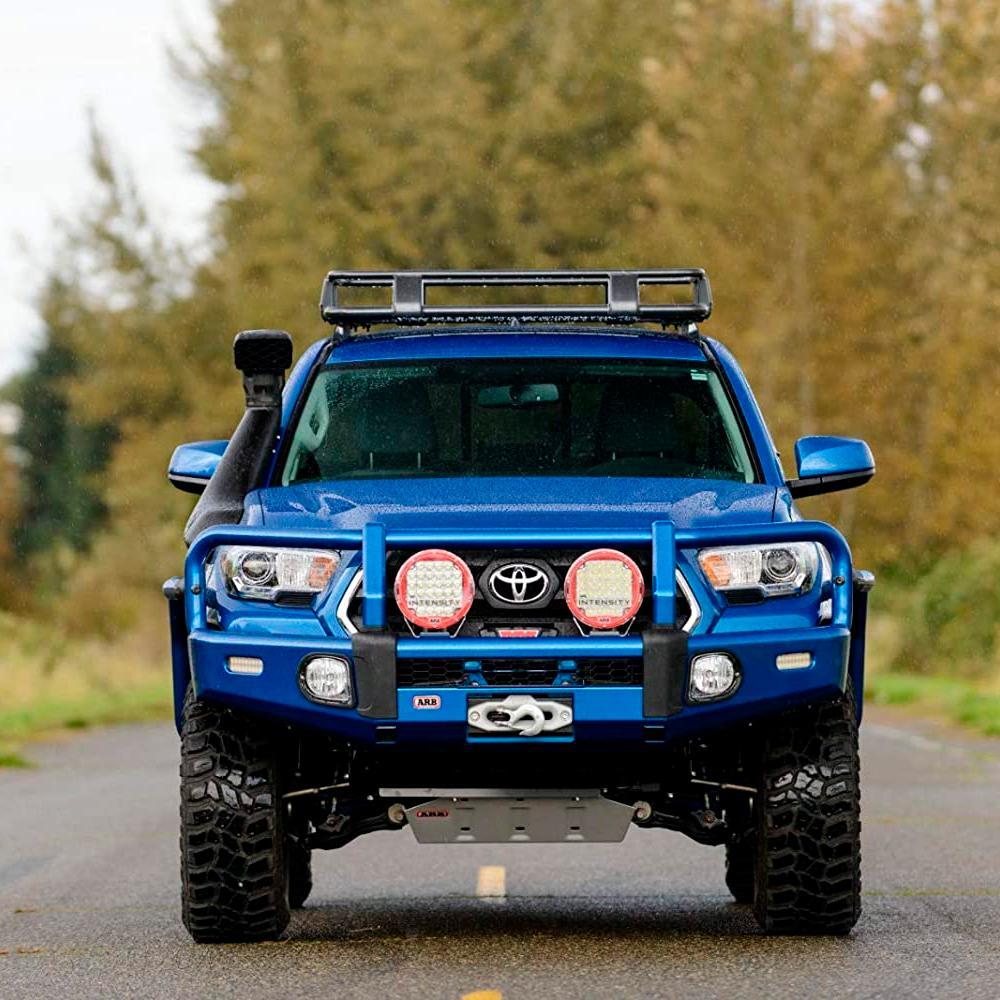 A blue Toyota Tacoma with an ARB Steel Roof Rack System for Jeep Wrangler 2018-2020 3813030KJL is driving down a country road, providing efficient cargo transportation.