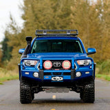 A blue Toyota Tacoma with an ARB Steel Roof Rack System for Jeep Wrangler 2018-2020 3813030KJL is driving down a country road, providing efficient cargo transportation.