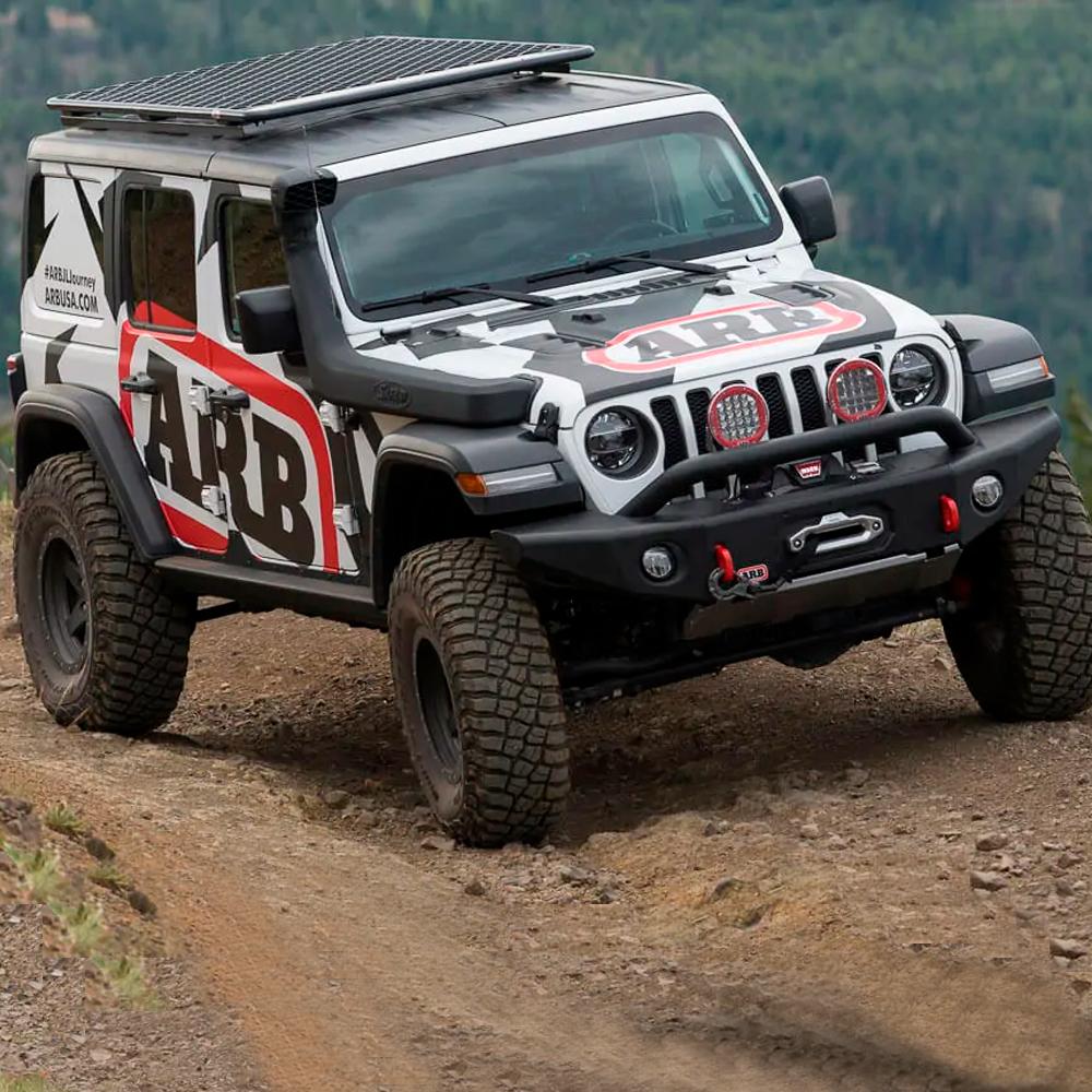 The ARB Alloy Flat Roof Rack 70” x 44” with Mesh Floor for Jeep Wrangler JL 2018-2019 is securely storing items in a fitting kit while driving down a dirt road.