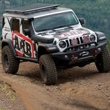The ARB Alloy Flat Roof Rack 70” x 44” with Mesh Floor for Jeep Wrangler JL 2018-2019 is securely storing items in a fitting kit while driving down a dirt road.