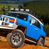 A blue Toyota FJ Cruiser with ARB roof racks is driving on a dirt road, equipped with the Roof Rack Components Toyota FJ Cruiser 2007-2014 ARB 3720110 high capacity mounting system.