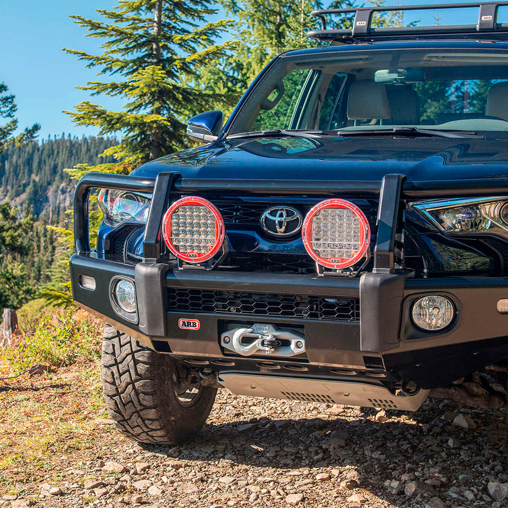 A black Toyota Tacoma, equipped with the ARB Under Vehicle Skid Plates System with kinetic (KDSS) 5421110 from ARB, is parked in a forest.