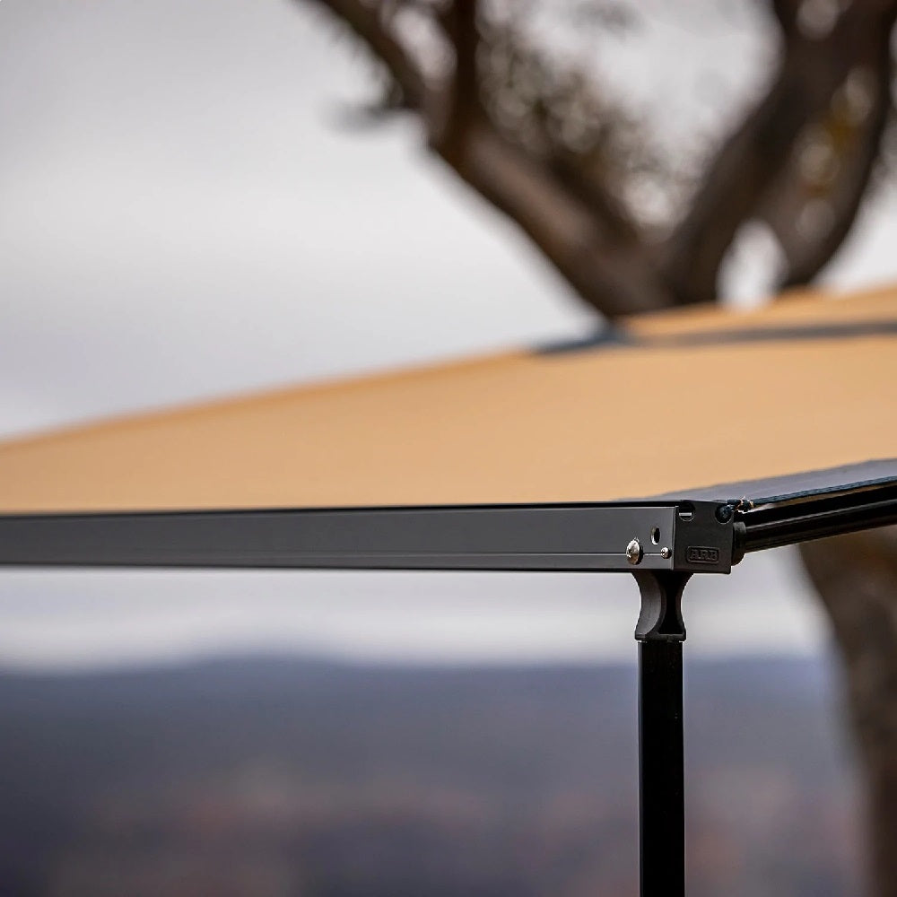 A close up of a tan ARB Touring Black Aluminum Awning with Light 814412A with a tree in the background.