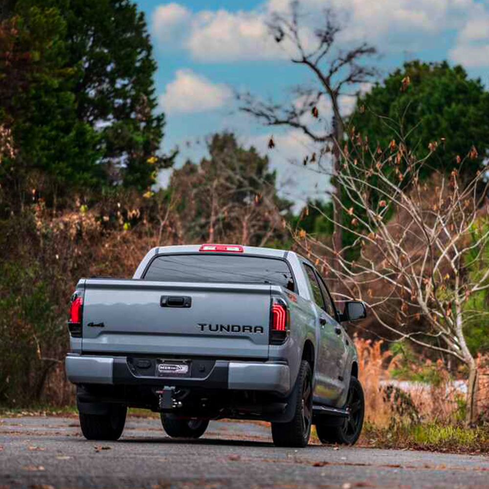 A gray truck with Morimoto Xb Led Tails LF705 for Toyota Tundra (2014 - 2021) aftermarket modifications is driving down a road with trees in the background.