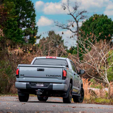 Load image into Gallery viewer, A gray truck with Morimoto Xb Led Tails LF705 for Toyota Tundra (2014 - 2021) aftermarket modifications is driving down a road with trees in the background.