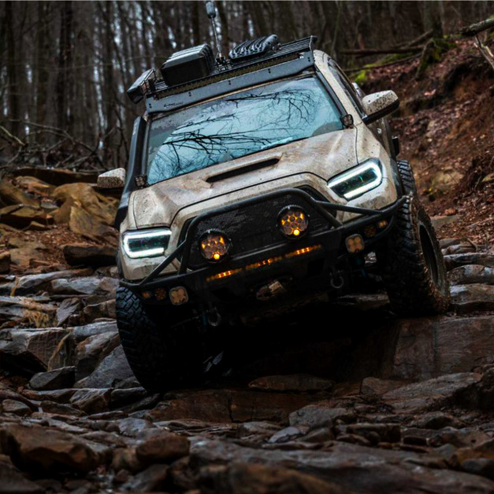 The 2020 Toyota Tacoma, equipped with aftermarket Morimoto XB Led Headlights LF530.2 ASM for Toyota Tacoma (2016 - 2022) from Morimoto, is driving through the woods.