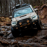The 2020 Toyota Tacoma, equipped with aftermarket Morimoto XB Led Headlights LF530.2 ASM for Toyota Tacoma (2016 - 2022) from Morimoto, is driving through the woods.