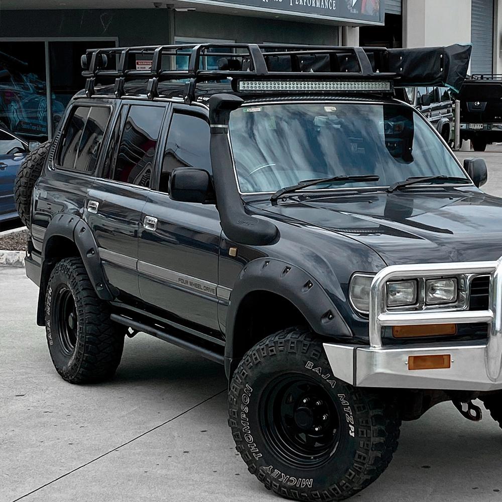 A high-quality ARB Steel Rack with Mesh Floor Kit 70" X 44" for Toyota Land Cruiser parked in front of a building, equipped with roof racks for increased load capacity.