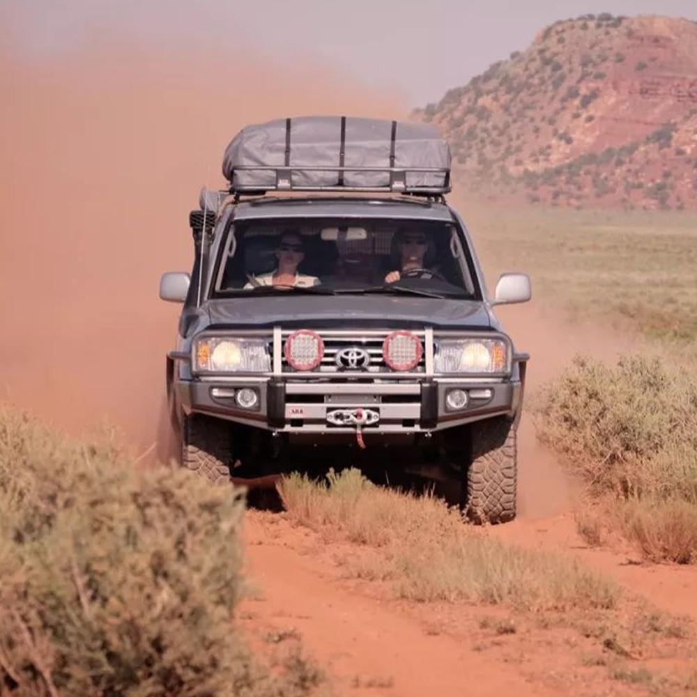 An ARB Roof Rack Fitting Kit 3713010 (87”x49”) for Toyota Landcruiser 100/105 Series and Lexus Lx470 driving down a dirt road, equipped with a fitting kit for secure storage and high capacity.