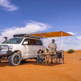 A Toyota Land Cruiser equipped with ARB roof racks and an ARB Touring Awning with Light 814409, perfect for camping in the waterproof tent in the desert.